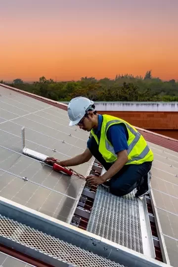 A man in a hard hat and high-vis vest kneels on a roof covered in solar panels, using a laptop and wires to install and fix the panels.